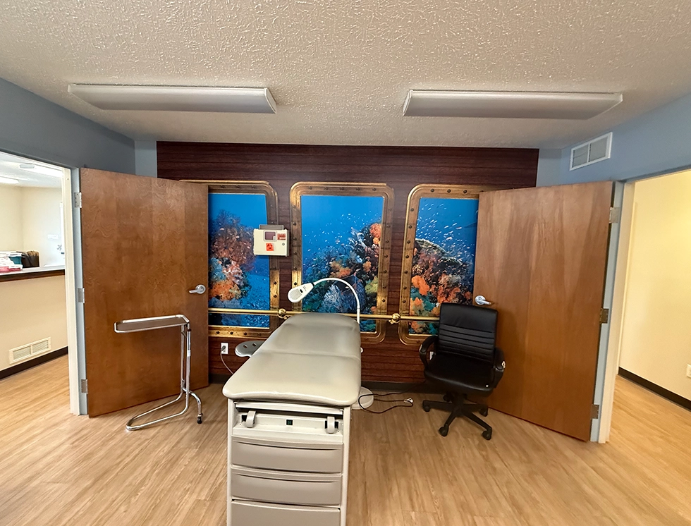 A treatment room featuring a padded exam table, a chair, and a vibrant underwater mural with coral and fish behind two wooden doors.